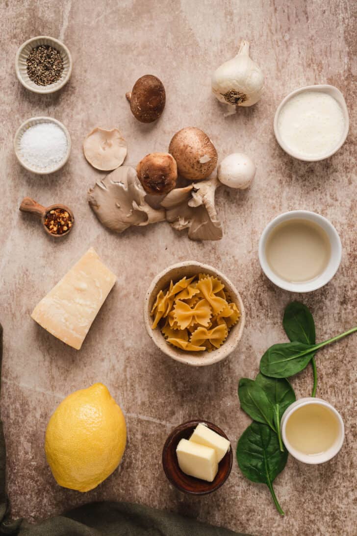 Ingredients laid out on a brown textured surface, including edible fungi, farfalle, cheese, spinach, lemon and spices.