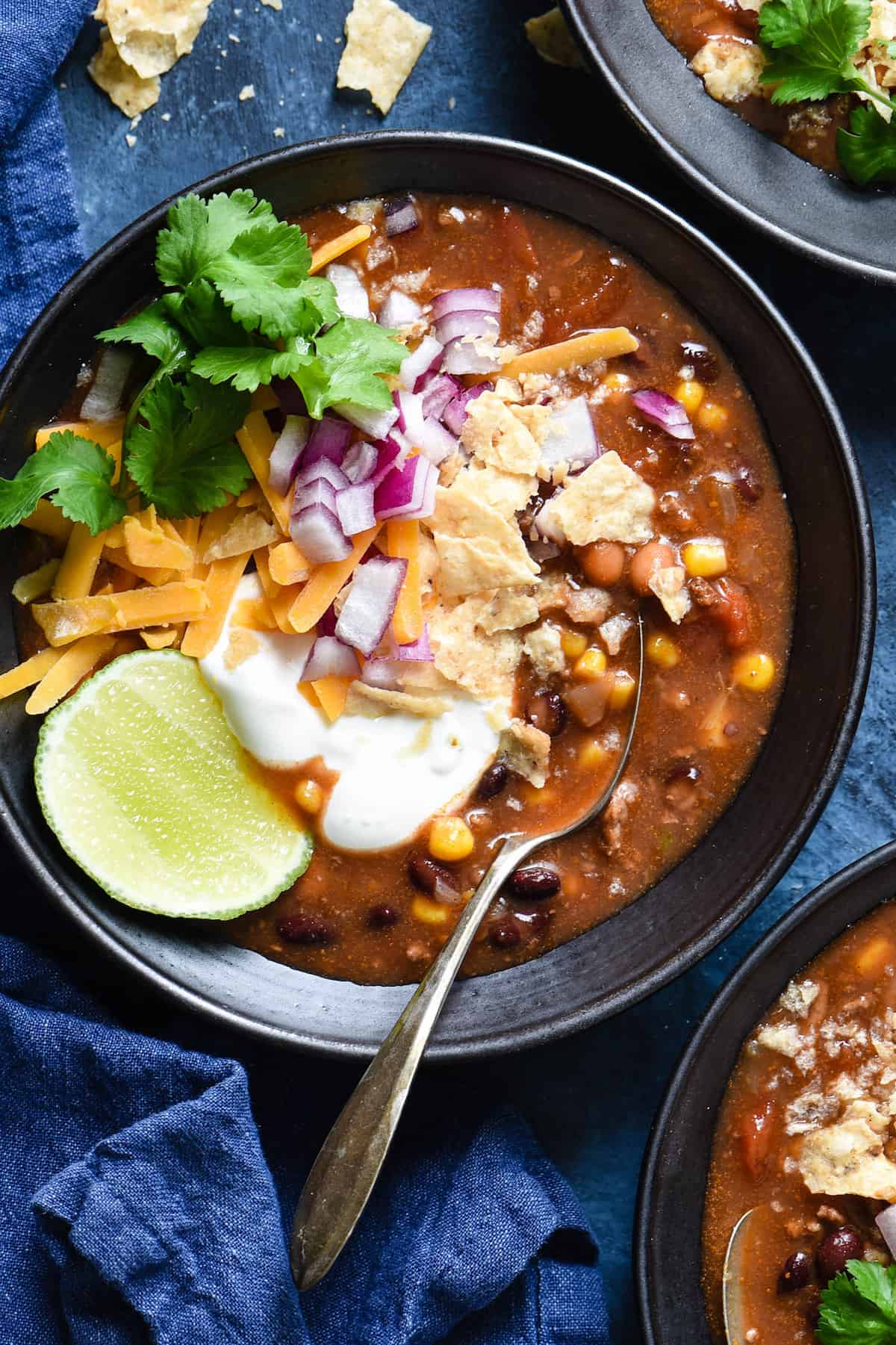 Black bowl filled with crockpot taco soup and garnished with sour cream, lime, cilantro, cheese, red onion and crushed tortilla chips.