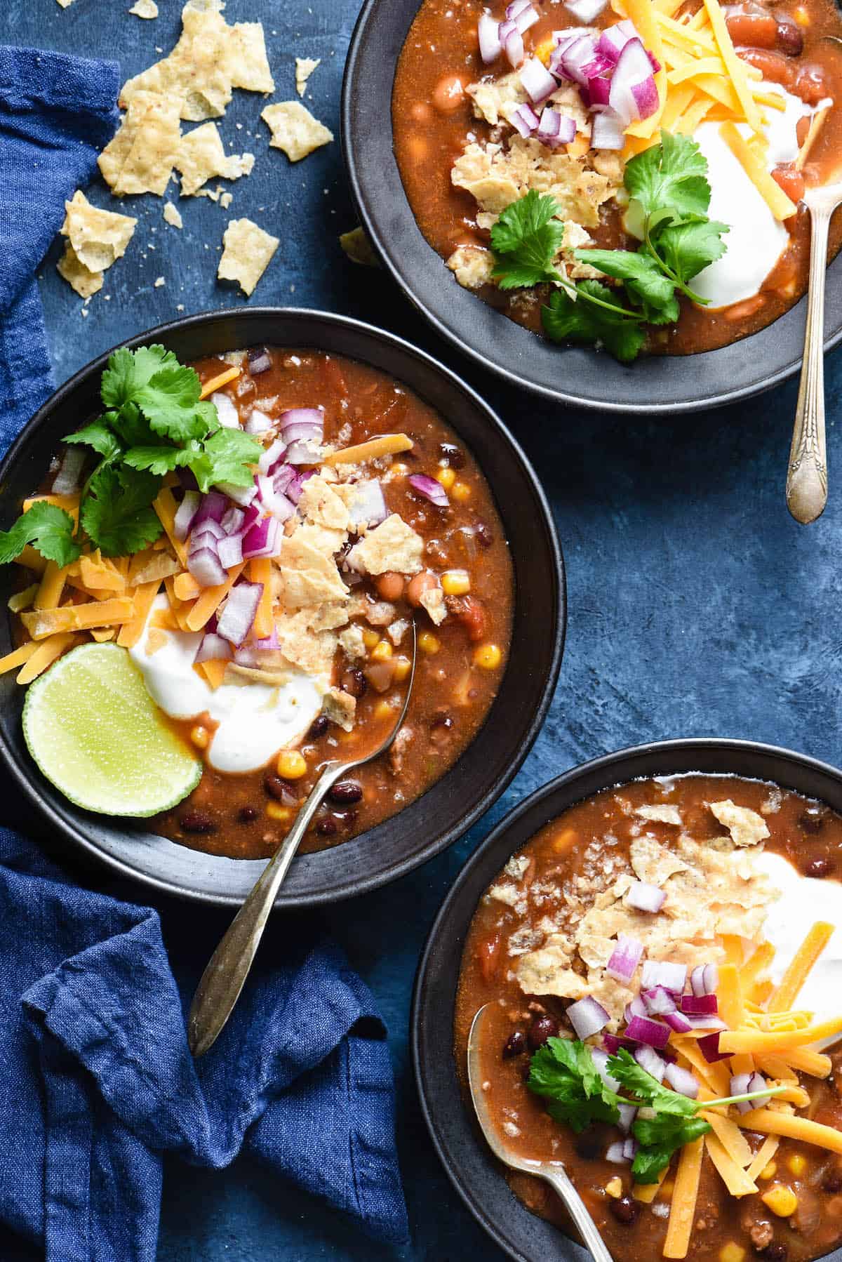 Three black bowls filled with a crockpot taco soup recipe and garnished with sour cream, lime, cilantro, cheese, red onion and crushed tortilla chips.