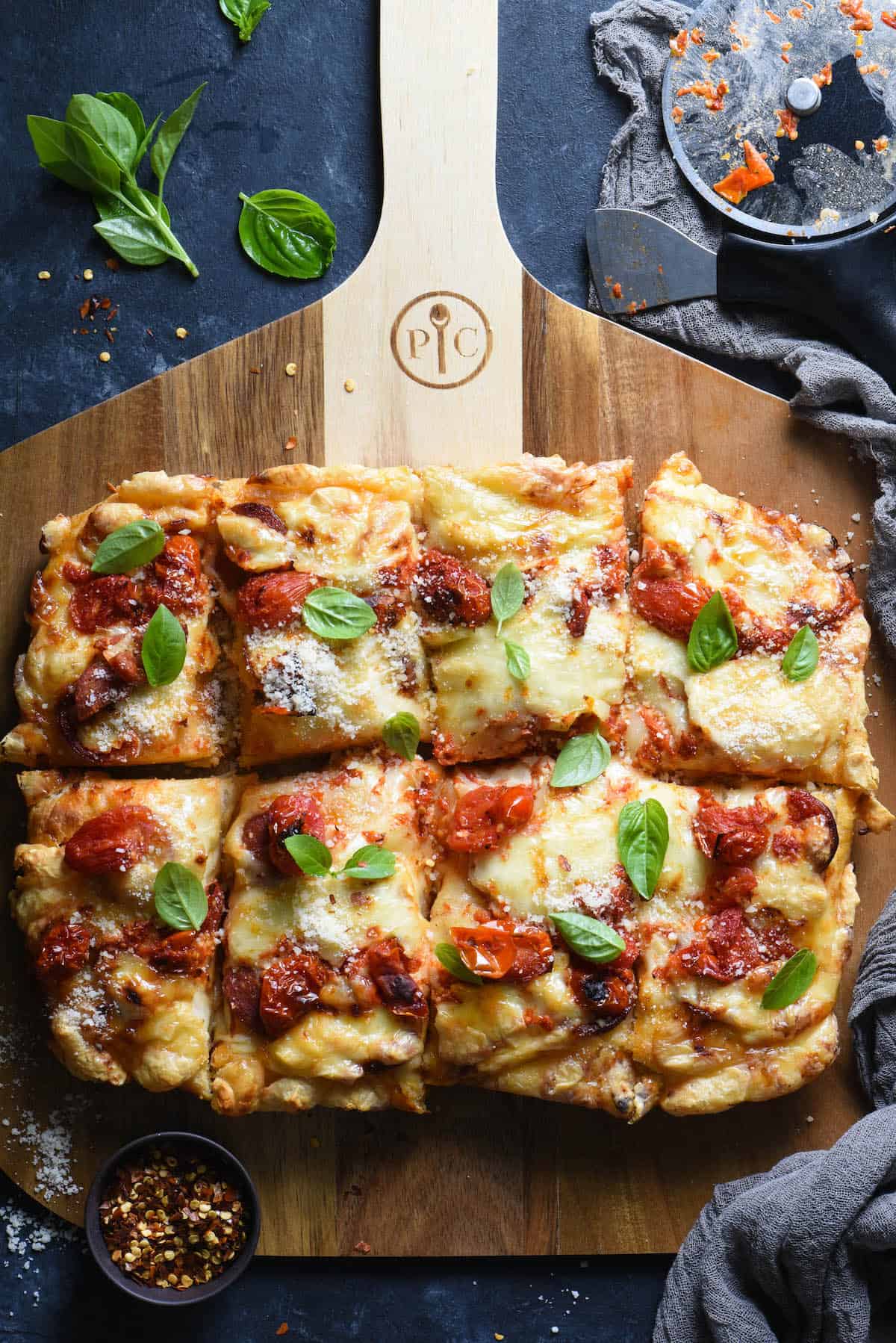 Rectangular deep dish pizza on wooden pizza board with pizza cutter and basil leaves on table.