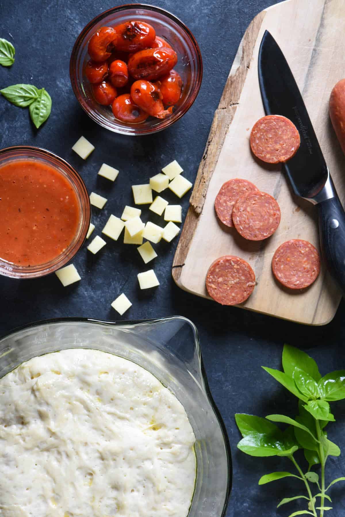 Dark tabletop with pepperoni, pizza dough, roasted tomatoes, basil and cubed cheese.