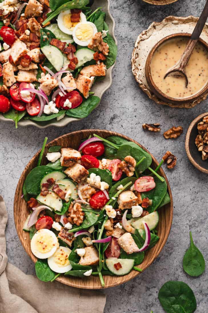 A wooden plate filled with spinach salad made with chicken, tomatoes, cucumbers, onion and eggs.