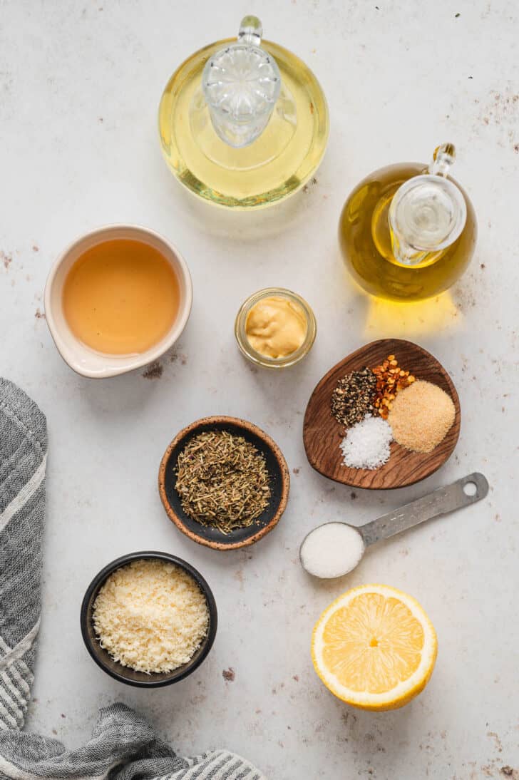 The ingredients for Italian salad dressing on a light surface, including oils, vinegar, herbs, spices, lemon, cheese and mustard.