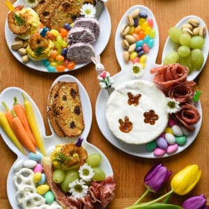 An Easter charcuterie board arranged on three white bunny head shaped plates. The plates are filled with charcuterie, cheeses, crackers, fruit, carrots, and candy.