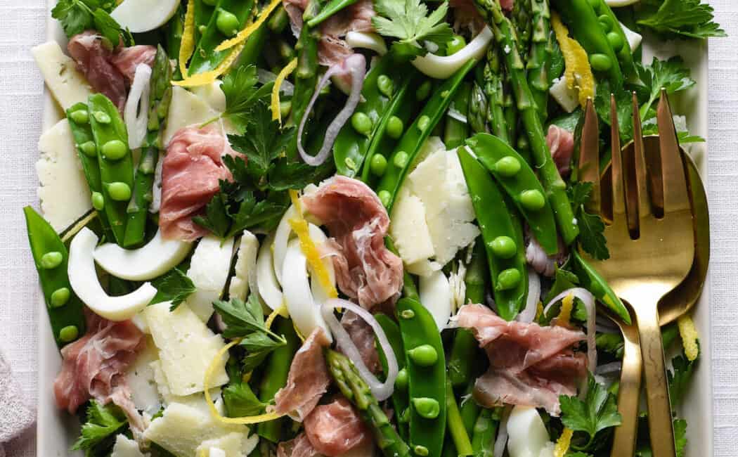 White platter filled with spring salad made with asparagus, snap peas, prosciutto, cheese and shallots.