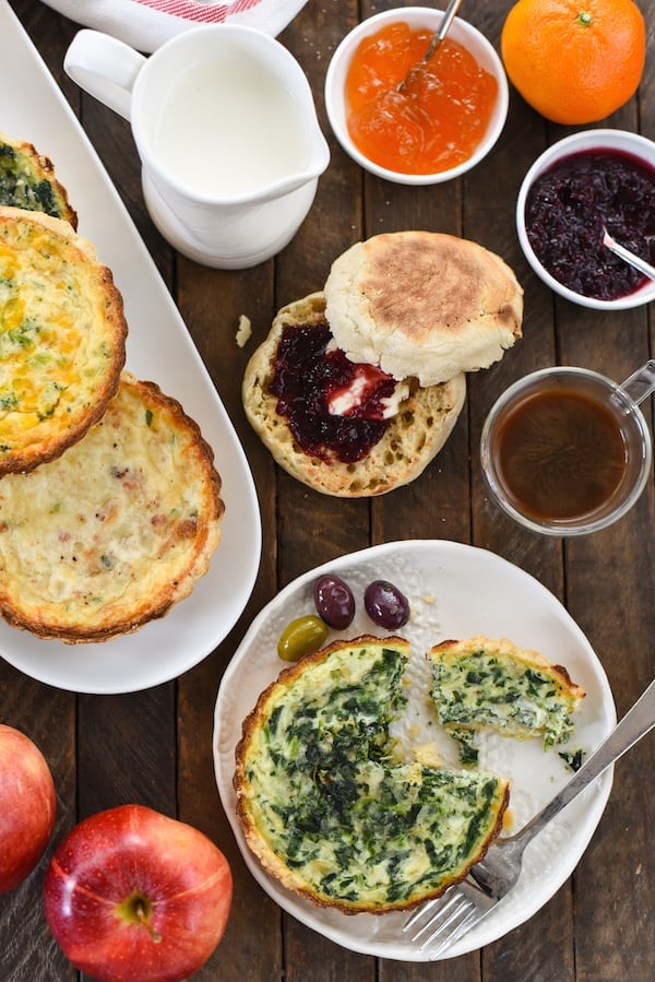 Overhead shot of brunch party table filled with mini quiches, olives, coffee, breakfast breads and spreads, salad and fruit.