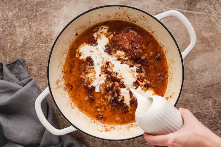 A white Dutch oven filled with a chunky red mixture, with a hand pouring cream over it.