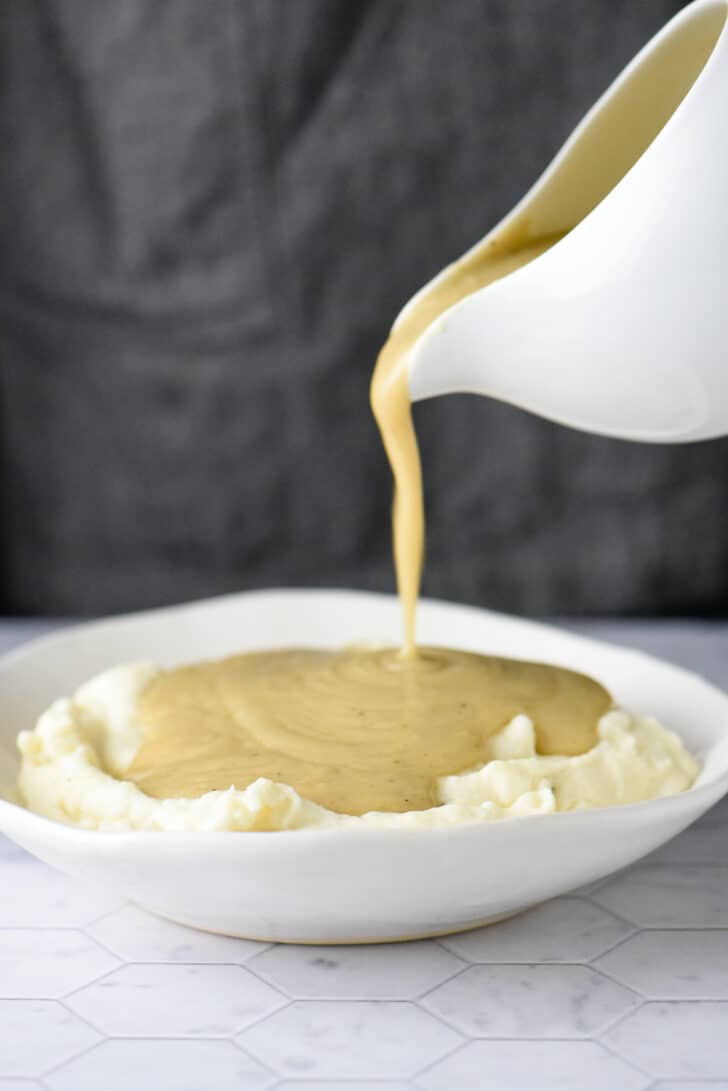 White gravy boat pouring gravy from scratch all over a bowl of mashed potatoes.