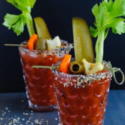 Two bloody mary cocktails with everything bagel rim salt and garnishes like celery, pickles and olives.