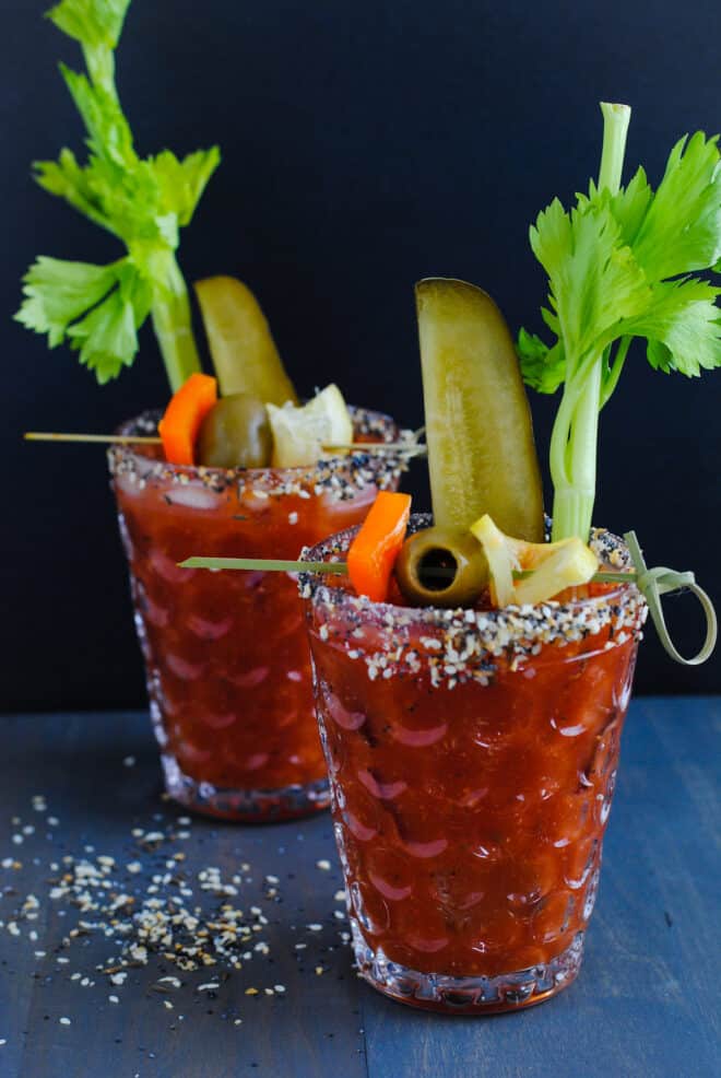 Two bloody mary cocktails with everything bagel rim salt and garnishes like celery, pickles and olives.