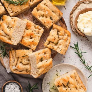 A loaf of focaccia bread cut into square pieces on a wooden cutting board and small plate.