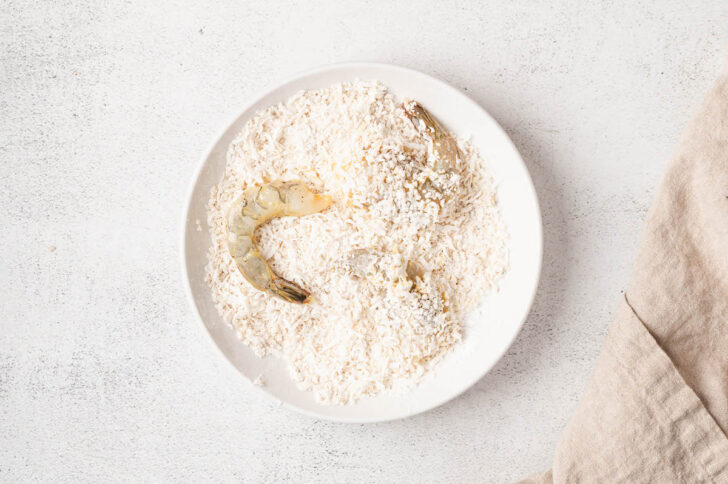 Prawns being crusted with a breading mixture in a white bowl.