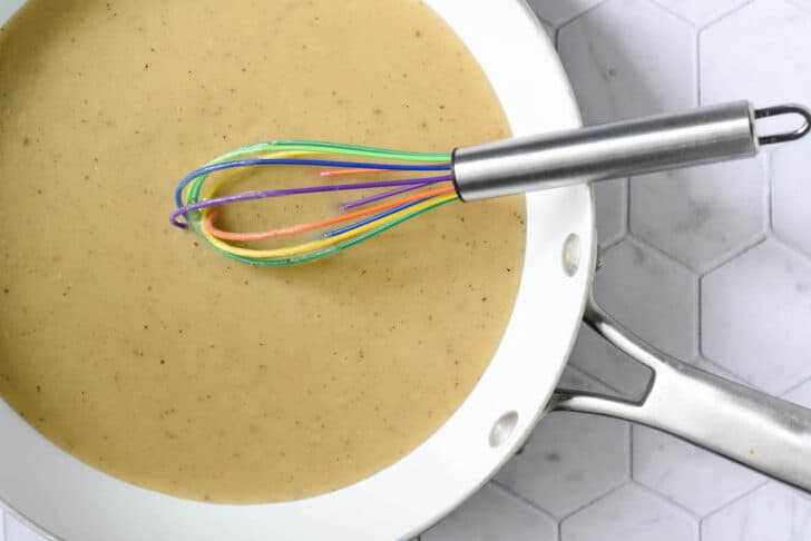 A white skillet filled with a light brown sauce flecked with spices, with a multicolored whisk.