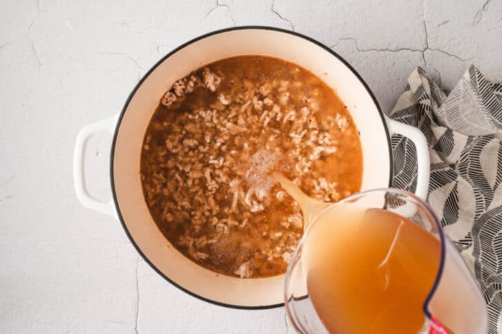 Broth being poured into a  white Dutch oven filled with cooked ground meat.