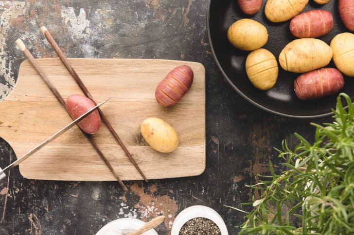 A knife cutting a potato crosswise on a cutting board. The potato is being held in place with two chopsticks.