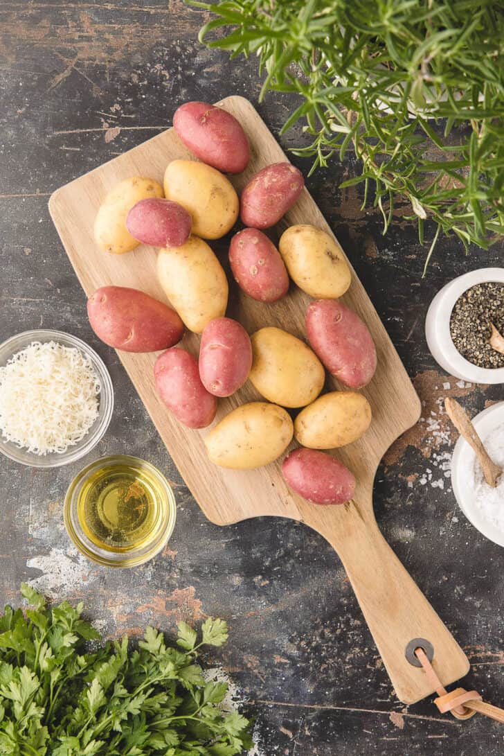 A cutting board filled with baby potatoes on a rustic surface, along with herbs, cheese and seasonings.