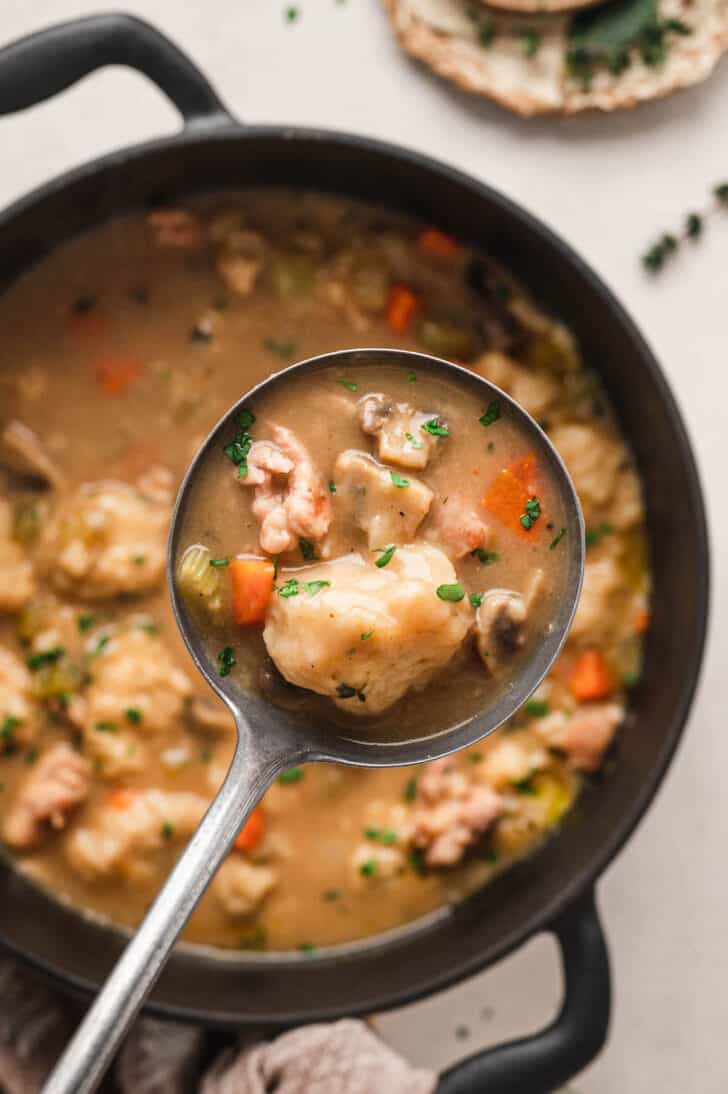 A black cast iron dutch oven filled with a homemade chicken and dumplings recipe, with a ladle spooning some out.