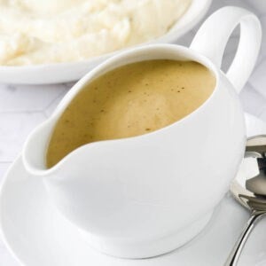 White ceramic boat filled with homemade gravy, with bowl of mashed potatoes in background.