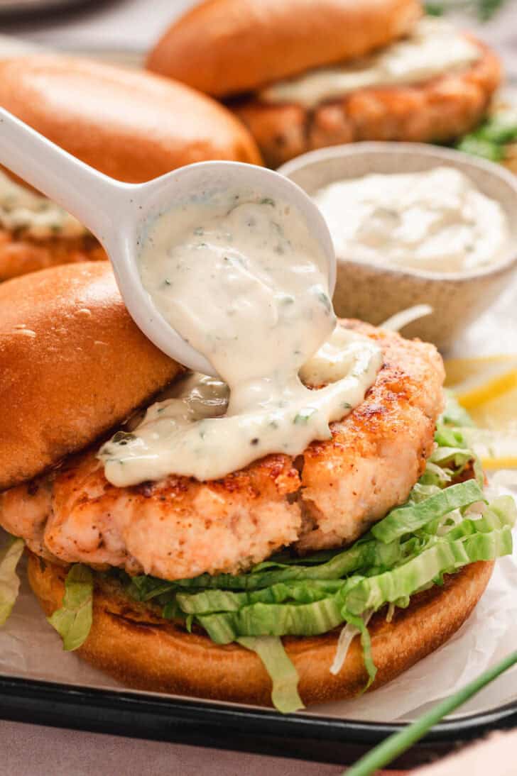 A spoon putting homemade tartar sauce on a salmon burger.