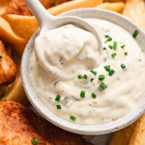 A spoon lifting a serving of a tartar sauce recipe from a white bowl, surrounded by fried fish and potatoes.