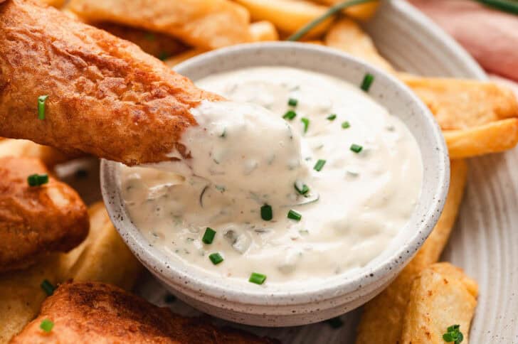 A piece of fried fish being dipped into a bowl of homemade tartar sauce.