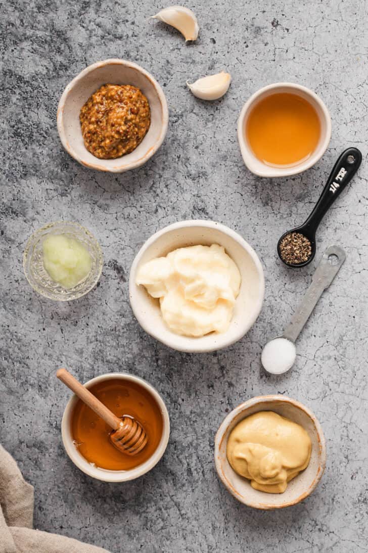 Ingredients on a textured gray surface, including mayonnaise, mustard, honey, spices and grated garlic and onion.