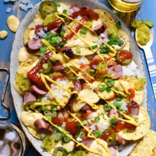 A large platter of nachos made with tortilla chips, cheese sauce, sliced hot dogs and pickled jalapenos.