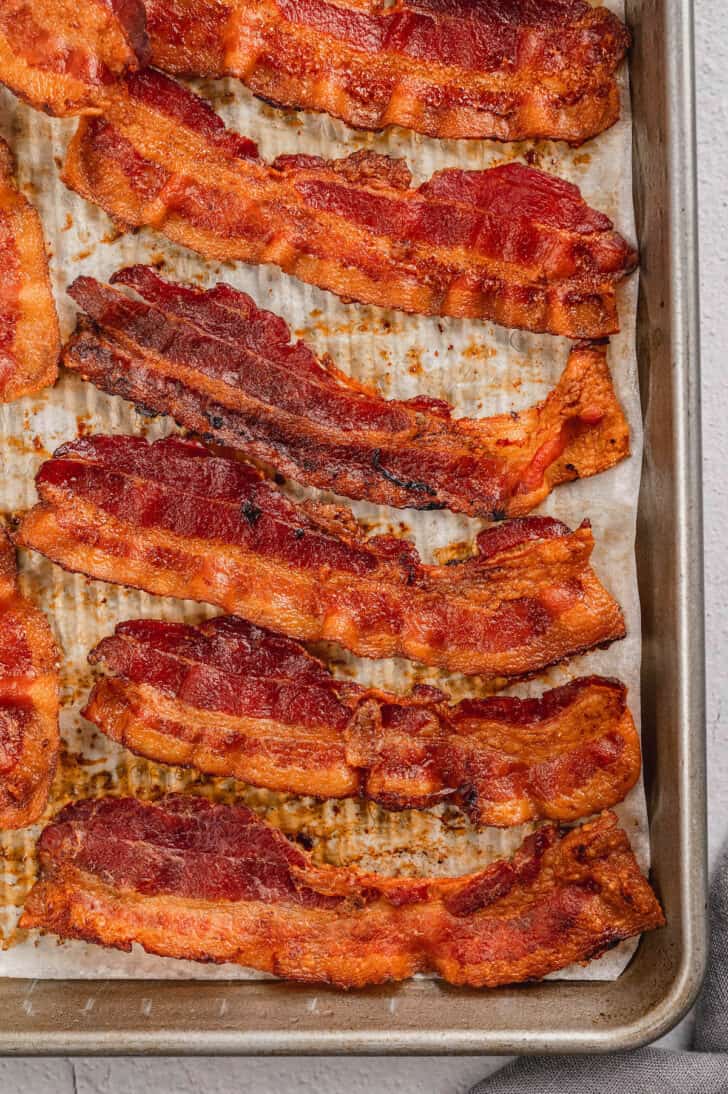 Bacon in oven lined up on a parchment paper lined baking pan.