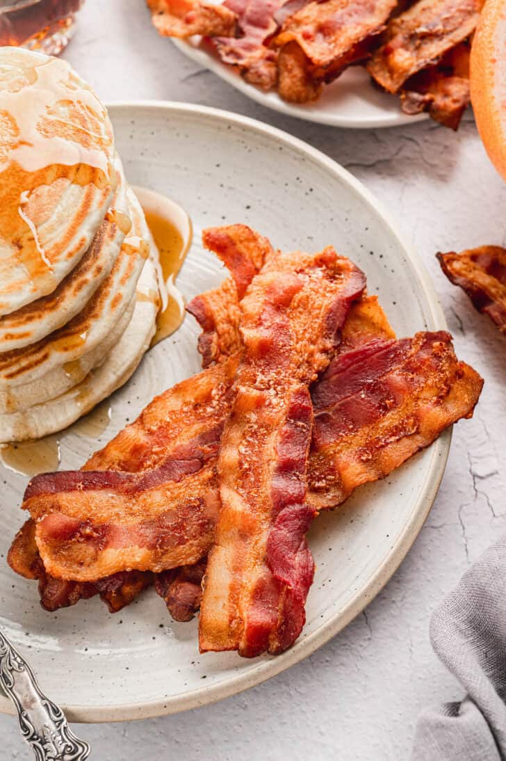 A speckled plate filled with a stack of pancakes with syrup, and the finished product of cooking bacon in oven.