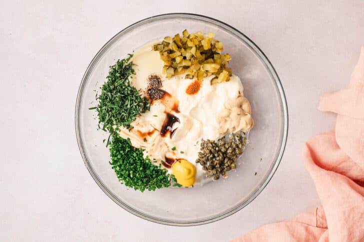 A glass bowl filled with various creamy ingredients, herbs, spices and condiments.