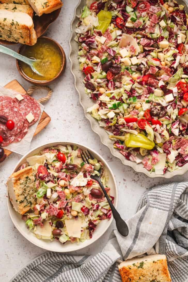 A scene showing chopped Italian salad on a large platter and a serving on a smaller plate, with garlic bread on the side.