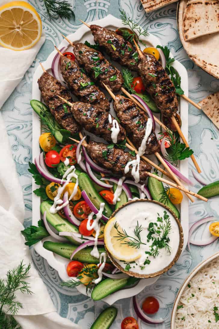 A large platter filled with kafta skewers, vegetables and a creamy dipping sauce.