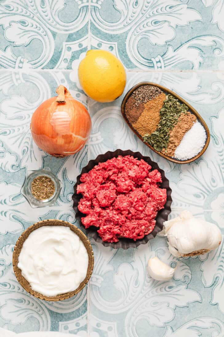 Ingredients on a light blue and white patterned surface, including ground meat, garlic, onion lemon, and dried herbs and spices.