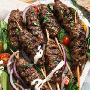 A large platter filled with kafta skewers, vegetables and a creamy dipping sauce.