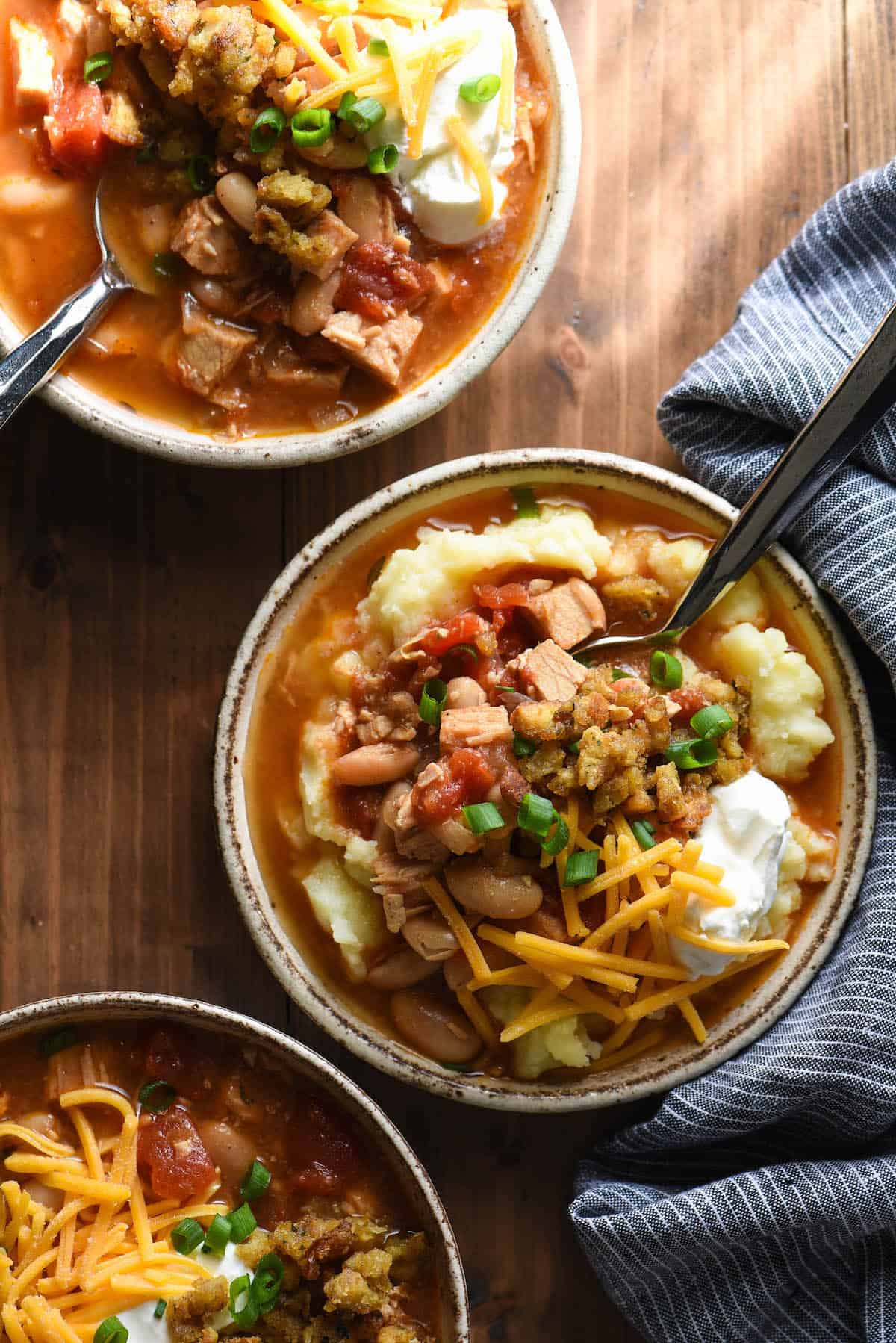 Three bowls of hearty chili garnished with cheese, sour cream and green onions, on wood table.