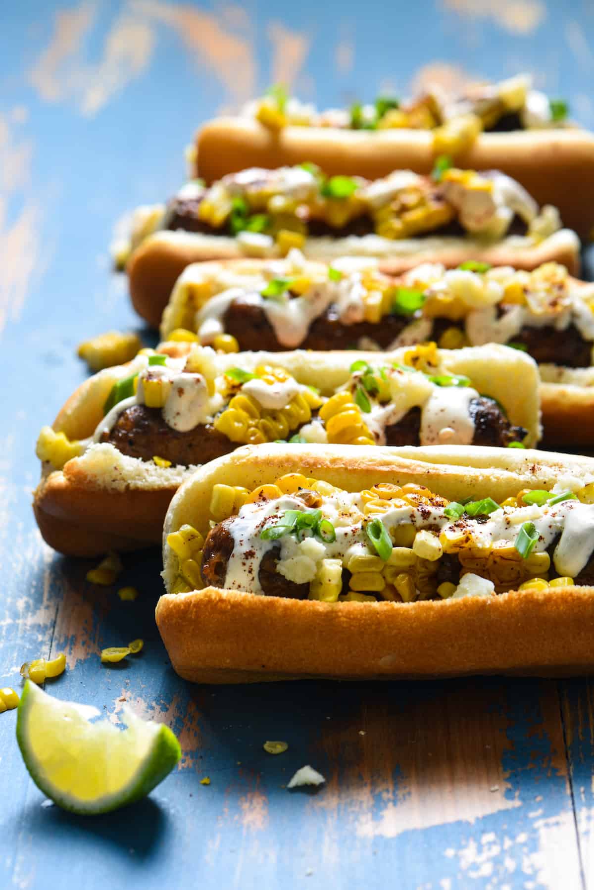 Inspired by elote, these Mexican Street Corn Brats are ideal for a summer celebration or easy weeknight meal! Spicy sausages are topped with fresh sweet corn, creamy lime sauce and tangy cheese. | foxeslovelemons.com