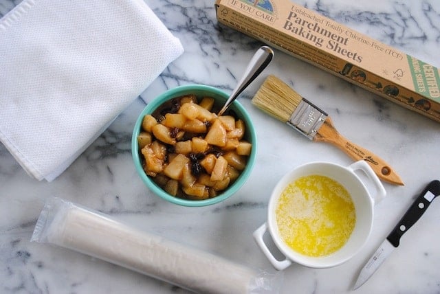 Mable countertop with white towel, bowl of cooked pears, bowl of melted butter, pastry brush, pairing knife, box of parchment baking sheets, and plastic-wrapped roll of phyllo dough.