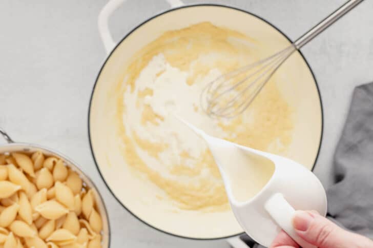 A hand pouring milk into a  white Dutch oven filled with roux.