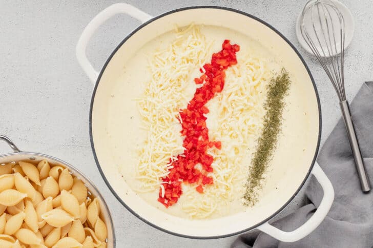 A white Dutch oven filled with a cream sauce topped shredded white cheese, chopped pimentos, and dried dill.