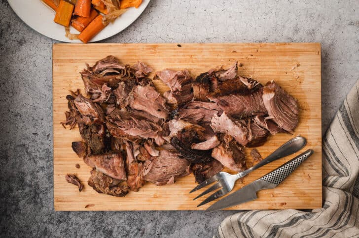 A crock pot pork roast being pulled apart on a wooden cutting board.