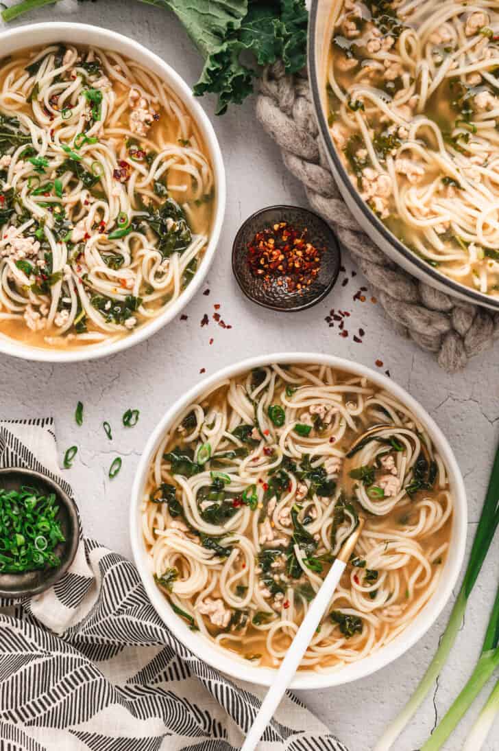 An overhead dinner table scene of shallow white bowls filled with ground pork soup with noodles.