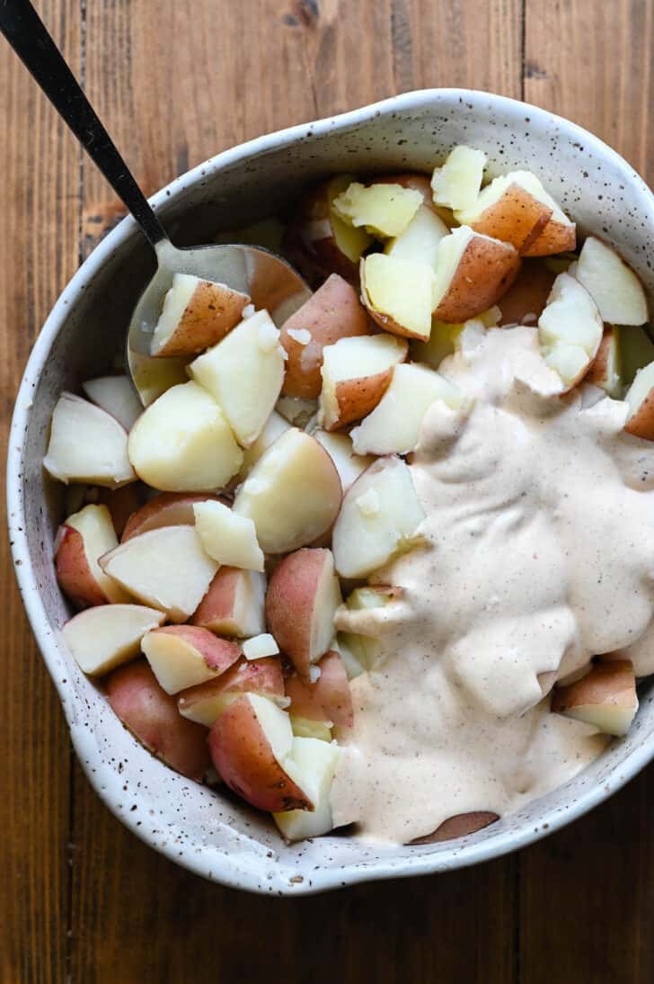 Cooked, chopped redskin potatoes in a speckled bowl with creamy dressing poured over half of the potatoes.