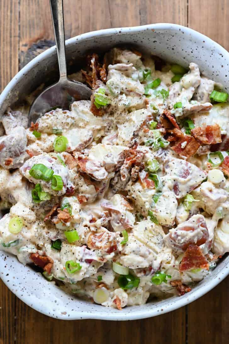 A speckled ceramic bowl filled with potato salad with bacon and green onions, with a spoon digging into the salad.