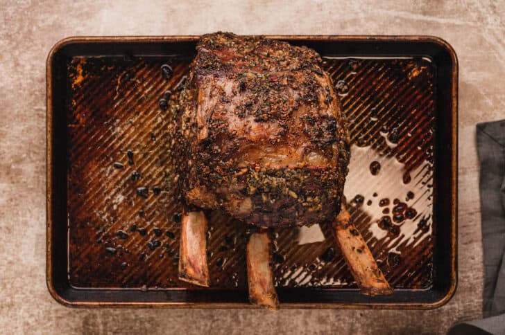 A rimmed baking pan topped with a cooked standing rib roast rubbed in herbs.