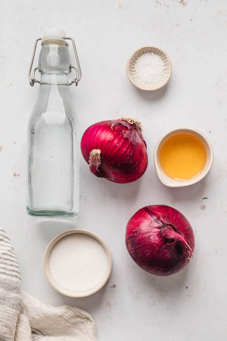 Ingredients on a white surface, including red onions, vinegar, sugar, salt and water.