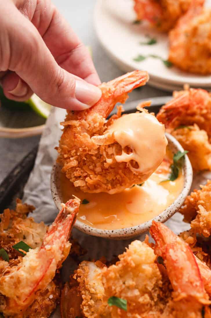 A hand dipping a panko-coconut crusted shrimp into an orange dipping sauce.