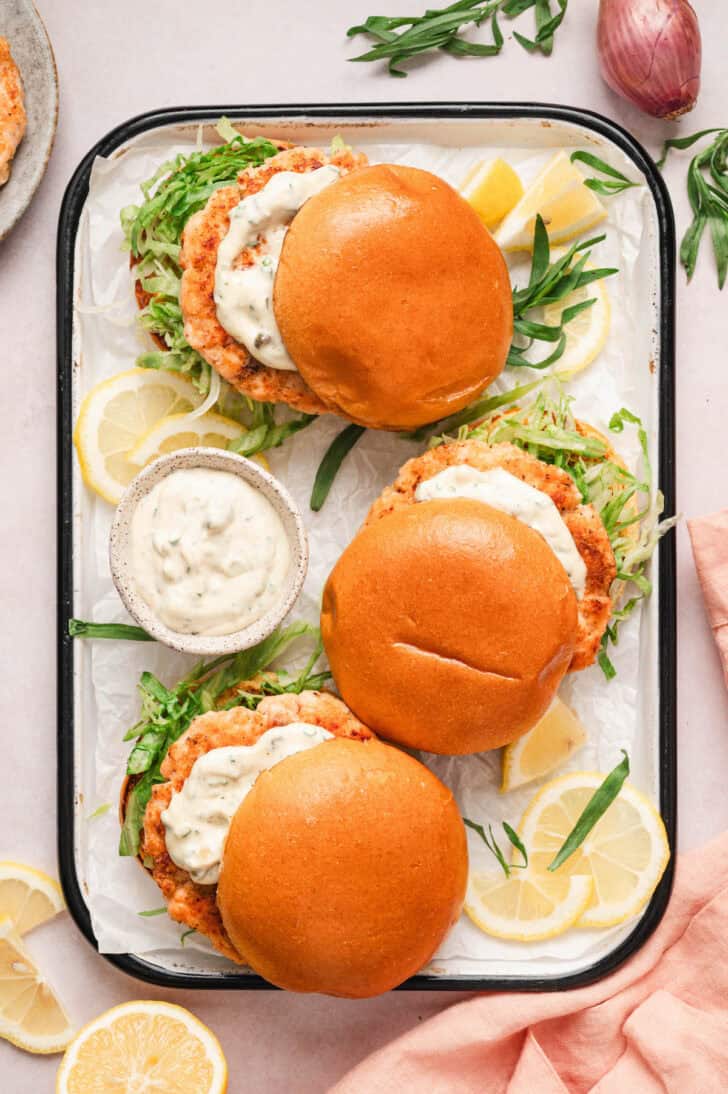 Three fresh salmon burgers on a tray, dressed with lettuce and tartar sauce.