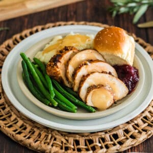 A turkey tenderloin recipe, sliced, on a plate with green beans, mashed potatoes and gravy, a roll and cranberry sauce.