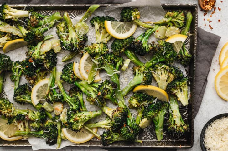 Roasted broccoli with lemon slices, shredded cheese and red pepper flakes on a textured baking pan.