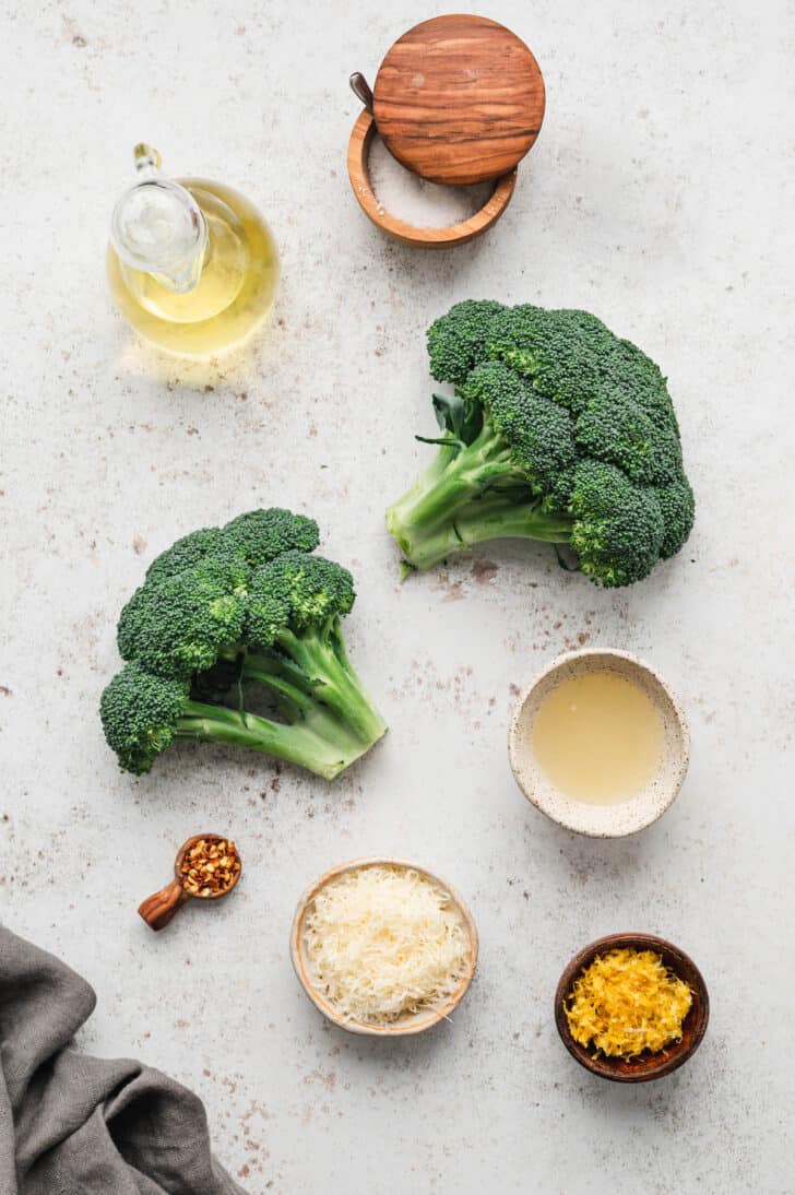 Ingredients laid out on a light surface, including fresh vegetables, olive oil, salt, lemon juice and zest, cheese and red pepper flakes.